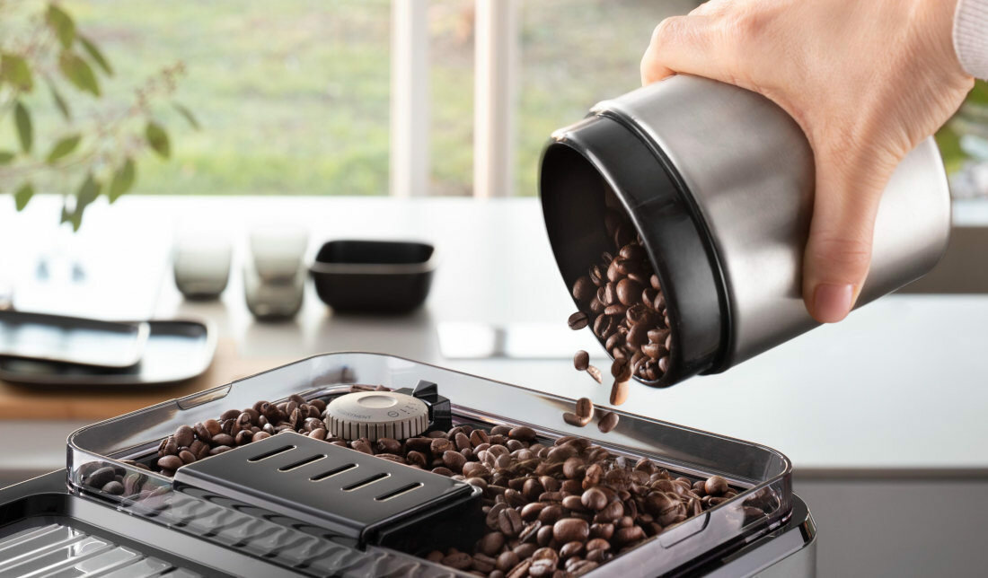 Zbliżenie na wsypywanie ziaren kawy do górnego zbiornika ekspresu DeLonghi Eletta Explore. Dłoń przechyla srebrny pojemnik, z którego lecą ziarna do otwartego młynka z widocznym pokrętłem regulacji. Tło jest jasne i rozmyte.