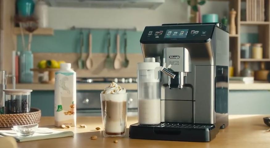 Ekspres DeLonghi Eletta Explore w aranżacji kuchennej z przygotowaną kawą mrożoną z bitą śmietaną. Obok urządzenia stoi napój migdałowy, co sugeruje możliwość przygotowania napojów roślinnych. W tle widać jasną, nowoczesną kuchnię z miętowymi akcentami.