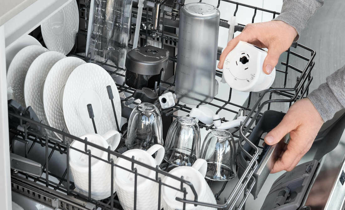 Mężczyzna wkłada elementy systemu LatteCrema ekspresu DeLonghi Eletta Explore do zmywarki. W koszu widoczne są karafki na mleko, pokrywy oraz domowe naczynia. Zdjęcie podkreśla, że akcesoria ekspresu można bezpiecznie myć mechanicznie.