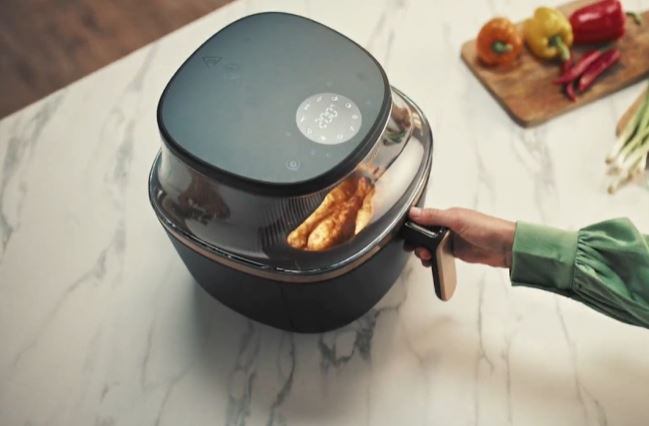 Рука открывает прозрачный ящик черной фритюрницы Philips Ovi NA322, открывая кусочки жареной курицы. На верхней панели фритюрницы есть цифровой дисплей с температурой «200°C» и значками программ приготовления. На светлом прилавке на заднем плане видны овощи на деревянной разделочной доске.