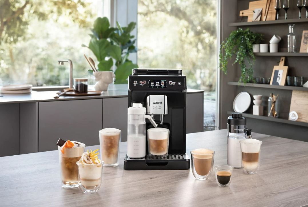 Automatyczny ekspres DeLonghi Eletta Explore w aranżacji kuchennej, otoczony różnorodnymi napojami kawowymi – od mrożonych deserów po klasyczne espresso. Na blacie widoczne są dwa systemy spieniania mleka: LatteCrema Cool i LatteCrema Hot. Tło stanowi jasna kuchnia z widokiem na ogród.