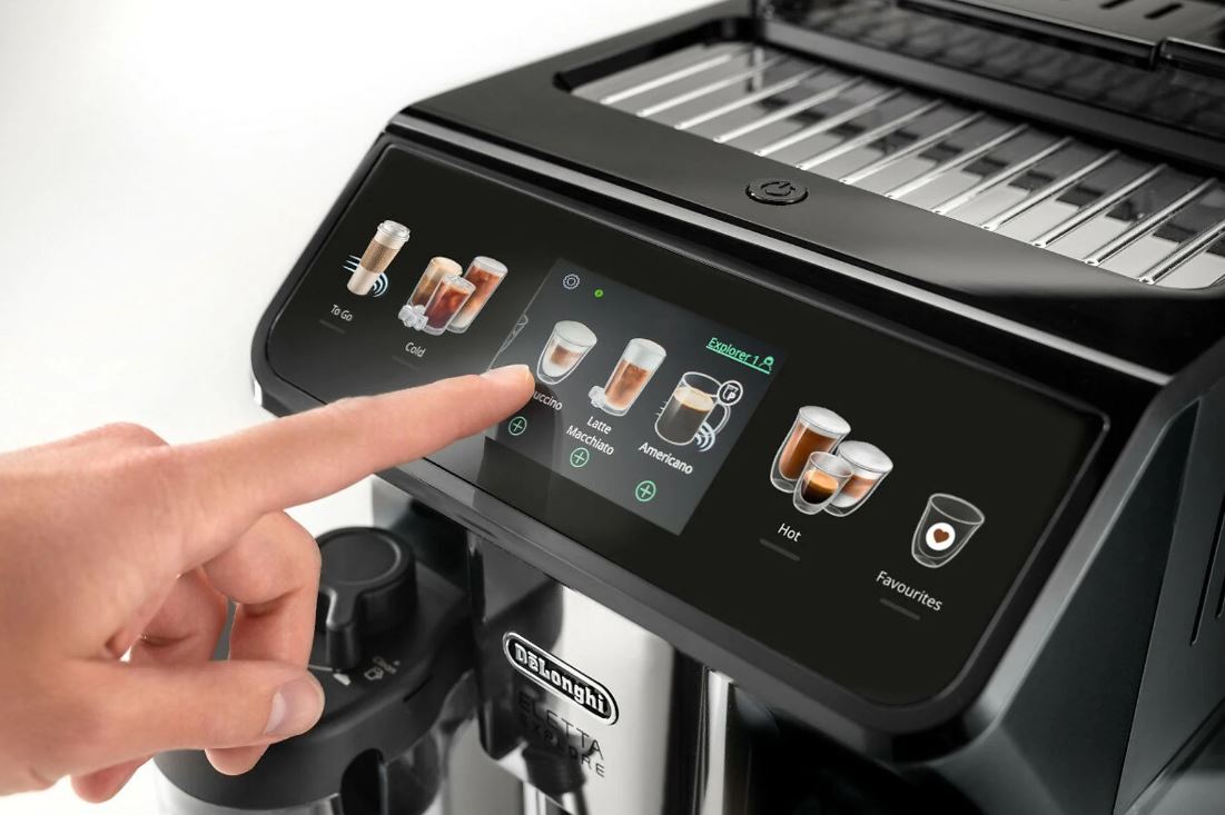 Zbliżenie na obsługę dotykowego panelu ekspresu DeLonghi Eletta Explore. Palec wybiera napój Cappuccino z szerokiej gamy dostępnych opcji wyświetlanych na kolorowym ekranie. Widoczne są kategorie napojów gorących, mrożonych oraz funkcja ulubionych.
